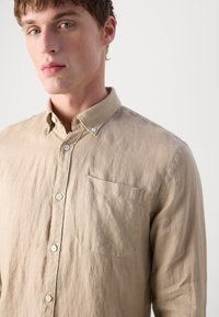 Camisa de linho bege com colarinho abotoado, bolso frontal e textura subtil. Apresenta um corte reto e detalhes em botão a condizer.