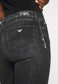 Vaqueros de denim negro con un diseño ajustado. Las características incluyen un parche con el logo en la cintura y un detalle bordado en el bolsillo.