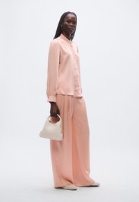 Lyserød silke bluse med knapper og lange ærmer, kombineret med rosa bukser med brede ben. Holder en lille, hvid håndtaske.