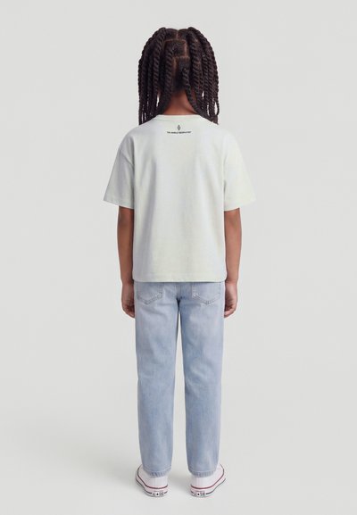 Παιδί με πλεγμένα στεφάνια που στέκεται γυρισμένο προς τα πίσω, φοράει άσπρο t-shirt, ανοιχτό μπλε τζιν και άσπρα αθλητικά παπούτσια σε απλό φόντο.