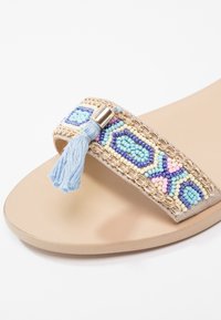 Sandale beige avec une bride décorée de perles multicolores, chaîne dorée et pompon bleu clair sur un fond uni.