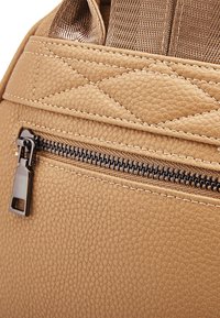 Sezione di zaino in finta pelle color tan con superficie testurizzata, dotato di una cerniera nera e di un tirante metallico, e dettaglio di cuciture trapuntate.