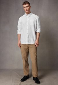 Chemise blanche à manches longues et à boutons avec une poche poitrine, portée avec un pantalon marron clair et des chaussures de ville noires sur un fond gris.