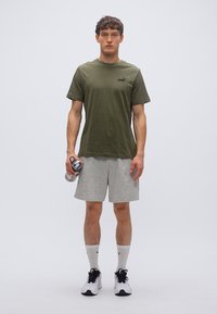 Πράσινο ελαιόχρωμο βαμβακερό t-shirt, γκρι σορτς, κρατώντας ένα μπουκάλι νερό. Λευκές κάλτσες με μαύρες λεπτομέρειες, μαύρα και λευκά αθλητικά παπούτσια. Απλό φόντο.