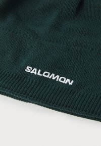 Zeleni pleteni beanie sa rebrastom teksturom i bijelim izvezenim "SALOMON" logotipom blizu donjeg ruba.