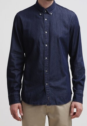 Mężczyzna ubrany w ciemnoniebieską dżinsową koszulę na guziki z długimi rękawami i beżowe spodnie, stojący na jednolitym tle.