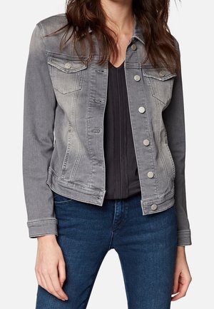 Kvinde iført en falmet grå denimjakke over en sort top og blå jeans, stående med afslappede arme mod en enkel baggrund.