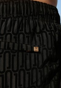 Crne kratke hlače s elastičnom trakom u struku, s tonovima u uzorku i zlatnim detaljem na džepu. Mekana tekstura s sjajnim završetkom.