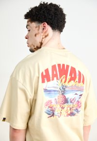 Beige t-paitti, jossa on suuri punainen graafinen kuva "HAWAII Aloha" trooppisine elementteineen, mukaan lukien ananas ja kukkakuviot selässä.