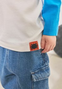 Korte mouwen witte t-shirt met blauwe lange mouwen, voorzien van een oranje en zwarte Spider-Man logo patch aan de zoom. Gecombineerd met denim broek.