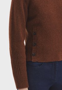 Brauner strukturierter Pullover mit geripptem Saum und Ärmelbündchen, ausgestattet mit vier dunklen Knöpfen an der Seite. Kombiniert mit dunkelblauen Jeans.