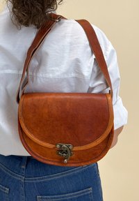 Osoba v modrých džínách a bílém tričku, která nese hnědou koženou crossbody tašku s kovovým zapínáním na zadní straně.
