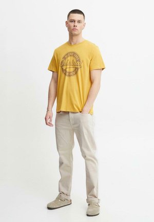 Camiseta de algodón amarilla mostaza con un estampado gráfico circular. Combinada con pantalones beige claros y zapatos de ante gris de tipo slip-on.