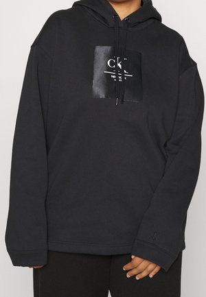 Persoon die een zwarte hoodie draagt met een glanzende vierkante patch aan de voorkant met het logo "CKL New York 97" en trekkoorden die naar beneden hangen.