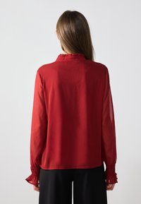 Rød bluse med rynket krage og lange, gjennomsiktige ermer, med rynkede mansjetter og en teksturert overflate. Vist fra baksiden.