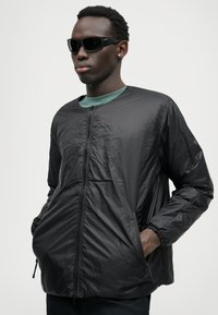 Giacca leggera nera con zip completa, polsini elasticizzati e due tasche laterali, indossata sopra una camicia verde, abbinata a occhiali da sole neri.