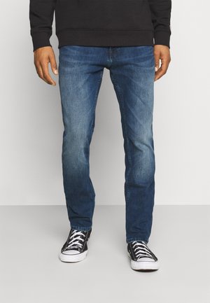 Mann trägt blaue Jeans, schwarzen Pullover und schwarze niedrige Turnschuhe mit weißen Schnürsenkeln, steht vor weißem Hintergrund.