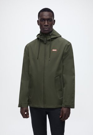 Mees, kes kannab oliivrohelist kapuutsiga jopet, millel on esitaskud ja lukuga esikülg, seisab ühtlase heleda tausta ees.