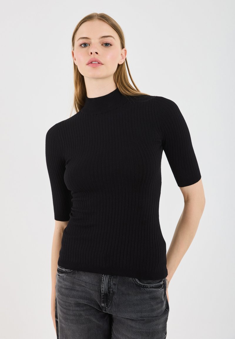 Melna rievota turtlenecka blūze ar īsām piedurknēm, ar pieguļošu siluetu un gludu tekstūru, apvienota ar tumši pelēkiem džinsiem.