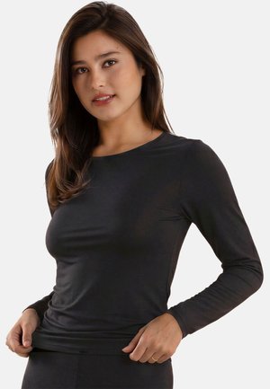 Young woman with long brown hair wearing a fitted black long-sleeve top, holding the bottom hem with both hands against a plain background.