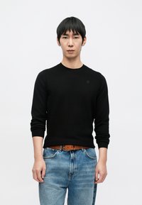 Juodas megztinis iš mezgimo su ilgomis rankovėmis, apvalia iškirpte ir subtiliu logotipu ant krūtinės, derinamas su mėlynais džinsais ir rudu diržu.
