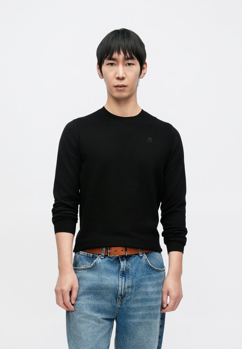 Juodas megztinis iš mezgimo su ilgomis rankovėmis, apvalia iškirpte ir subtiliu logotipu ant krūtinės, derinamas su mėlynais džinsais ir rudu diržu.