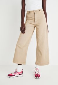 Béžové široké kalhoty vyrobené z bavlny, s předním zapínáním na knoflík. V kombinaci s růžovými a vínovými teniskami a bílými ponožkami.