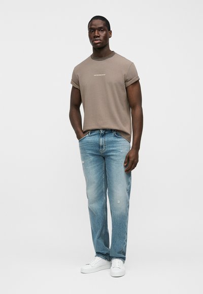 Καφέ βαμβακερό t-shirt με γυρισμένα μανίκια, που έχει λευκό λογότυπο, συνδυασμένο με ανοιχτό μπλε τζιν με φθορές και λευκά αθλητικά παπούτσια.