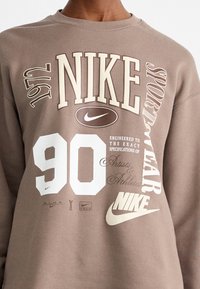 Brūna kapuce ar dažādiem baltiem un krēmveida grafikiem, tostarp "Nike", "Sportwear" un "90", kā arī apaļš Nike logo.