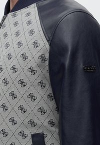 Veste en tissu gris à l'avant avec motif répétitif de logo noir, manches en cuir bleu marine et badge métallique « GUESS Est. 1981 » sur la manche.