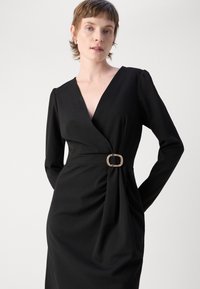 Robe noire portefeuille à manches longues, décolleté en V profond, plis centraux, et une boucle de ceinture rectangulaire dorée à la taille. Texture lisse, design ajusté.