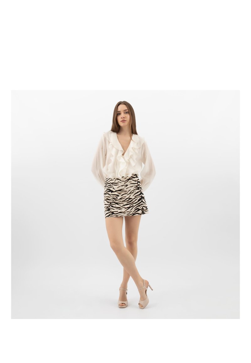 Jeune femme debout, jambes croisées, portant un chemisier blanc à volants, une mini-jupe à imprimé zèbre et des sandales beige à talons hauts, sur un fond blanc.