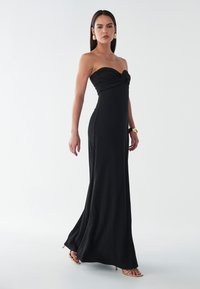 Melns bezplecu kleita ar pieguļošu siluetu, pagrieztu korpusa detaļu, plūstošu grīdas garuma svārku un gludu, teksturētu audumu.