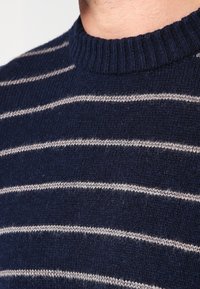 Námořnický svetr s horizontálními béžovými pruhy, s ribbovaným kulatým výstřihem a měkkou vlněnou texturou. Detailní pohled, zdůrazňující texturu látky.