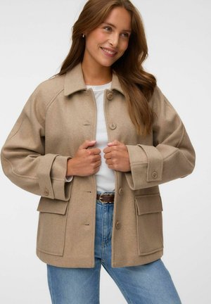 Frau lächelt, trägt eine beige, geknöpfte Jacke mit großen Taschen, ein weißes Hemd, blaue Jeans und einen braunen Gürtel, steht vor weißem Hintergrund.