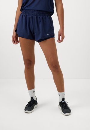 Osoba ubrana w granatowe sportowe spodenki i top, białe skarpety Nike oraz czarne buty do biegania, stojąca na prostym białym tle.