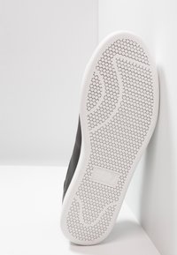 Sula av en svart sneaker med vit texturerad sula och EA7 Emporio Armani-logotypen i mitten, lutande mot en vit hörnvägg.