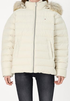 Femme portant une doudoune crème zippée avec capuche bordée de fourrure et poches latérales, associée à un jean skinny foncé.