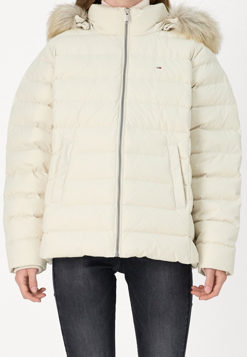 Femme portant une doudoune crème zippée avec capuche bordée de fourrure et poches latérales, associée à un jean skinny foncé.