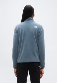 Ανδρικό μπλε fleece μπουφάν με ψηλό γιακά, απαλό ύφασμα και λεπτομέρεια λογοτύπου στην πλάτη, συνδυασμένο με μαύρο παντελόνι με διακοσμητικές τσέπες.