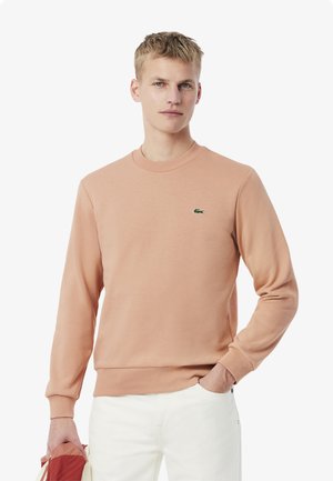 Sudadera de melocotón claro con mangas largas, cuello y puños acanalados, que presenta un pequeño logotipo verde en el pecho. Tejido de mezcla de algodón suave.