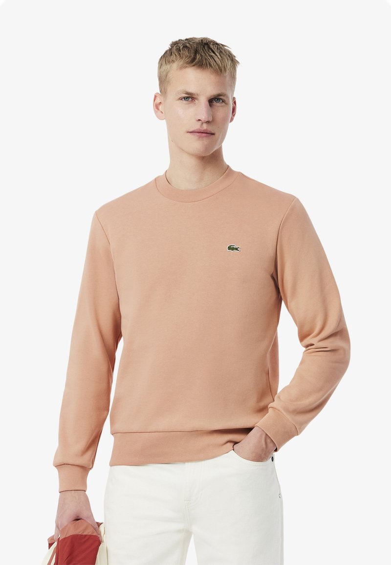 Sudadera de melocotón claro con mangas largas, cuello y puños acanalados, que presenta un pequeño logotipo verde en el pecho. Tejido de mezcla de algodón suave.