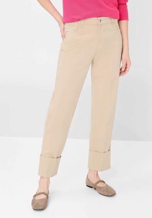 Personne portant un pantalon beige à jambes larges avec de grands revers, un haut rose vif et des ballerines beiges, debout devant un fond uni clair.