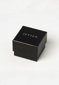 Sort firkantet smykkeskrin med tekstureret finish og et sølvpræget logo "JETTE" på topfladen, med hængslet låg.