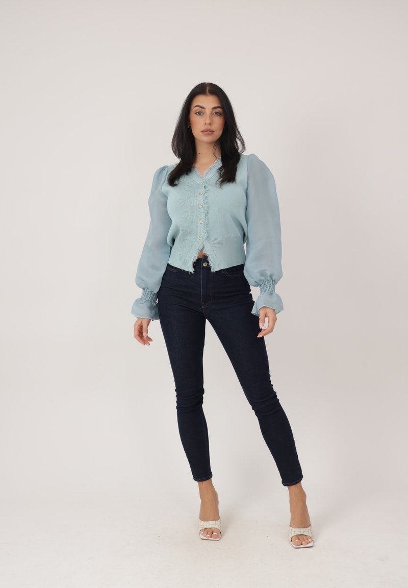 Mujer de pie contra un fondo liso, vistiendo una blusa azul claro con botones y mangas abullonadas, jeans ajustados oscuros y tacones blancos abiertos en la punta.