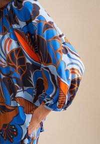 Blusa multicolore con motivo floreale in blu, arancione e marrone, con vestibilità ampia e maniche a sbuffo. Tessuto morbido e liscia.