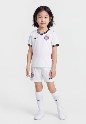 Copil tânăr în picioare, purtând echipament de fotbal alb al Angliei cu logo-uri Nike, inclusiv tricou, pantaloni scurți, șosete lungi și adidași albi, pe un fundal simplu.
