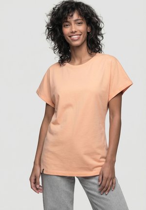 Lächelnde Frau mit lockigem Haar, die ein pfirsichfarbenes T-Shirt mit kurzen Ärmeln und graue Hose trägt, vor einem schlichten hellen Hintergrund stehend.