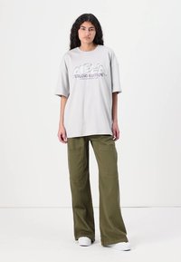 T-shirt oversize gris avec un imprimé graphique portant l'inscription "ALPHA STUDIO EDITION", associé à un pantalon cargo vert ample. Chaussures blanches et fond uni.