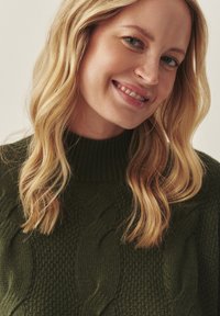 Tumši zaļš adīts džemperis ar augstu apkakli, kam ir teksturēti kārtojumi un mīksta, blīva auduma struktūra. Garas, viļņotas matu šķipsnas redzamas.